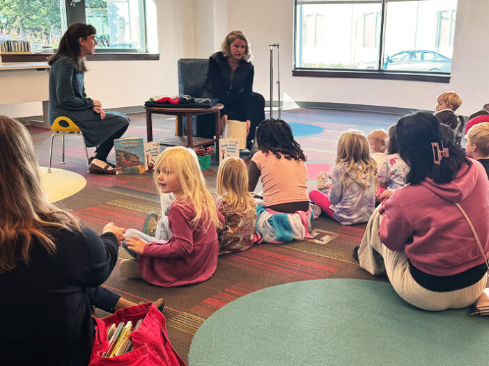 Photo of Storytime at the Library with children sitting on the floor and watching a librarian and musician interact.